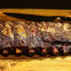Two-thirds of a rack of Pen’s House ribs, glazed and sliced with bones disappearing out of frame and a large kitchen knife
