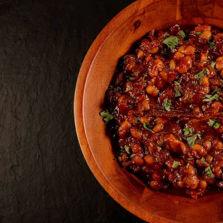 Wooden bowl filled with Pendragon's Smokehouse in house made smokey BBQ baked beans on a dark background