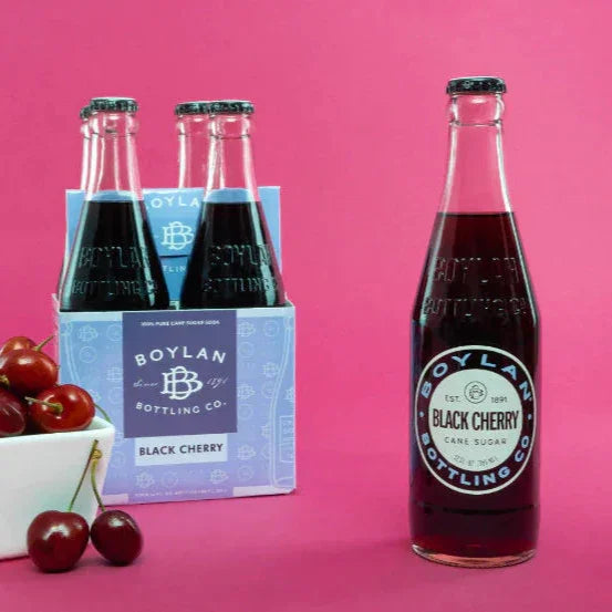 A glass bottle of Boylan Black Cherry soda with a pack of four bottles visible in the background, alongside a bowl of cherries. The foreground bottle is prominently displayed with a label that includes the brand name and the flavor 'Black Cherry'.