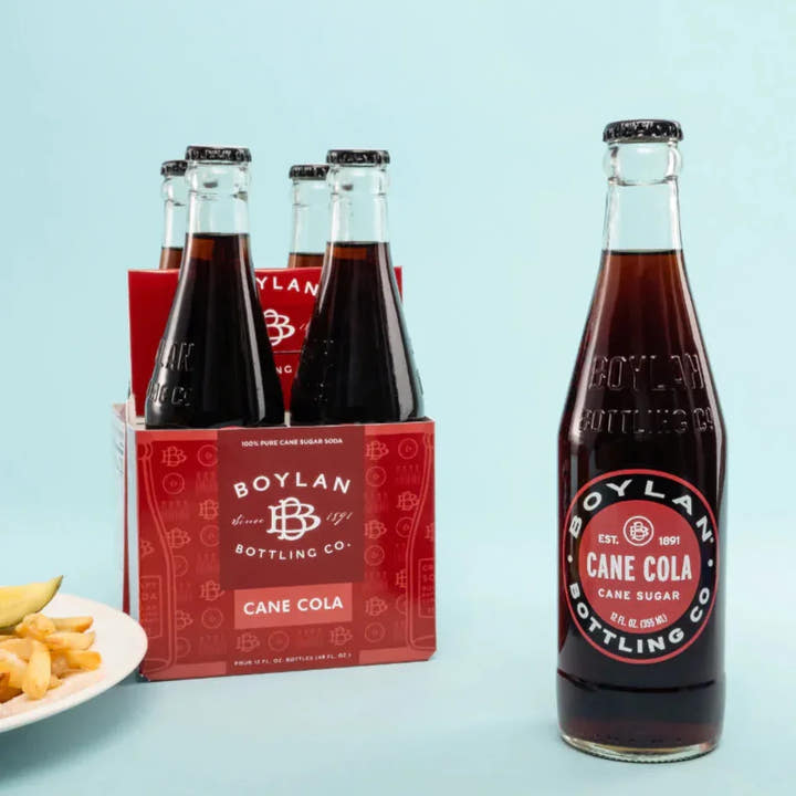 A four-pack of Boylan Cane Cola bottles next to a single bottle displayed in front, with a plate of fries in the background.