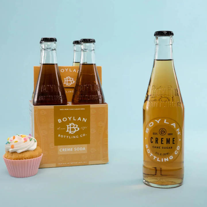 A 12oz bottle of Boylan Crème Soda with a pack of four bottles in the background, alongside a cupcake. All items are displayed against a light blue background.