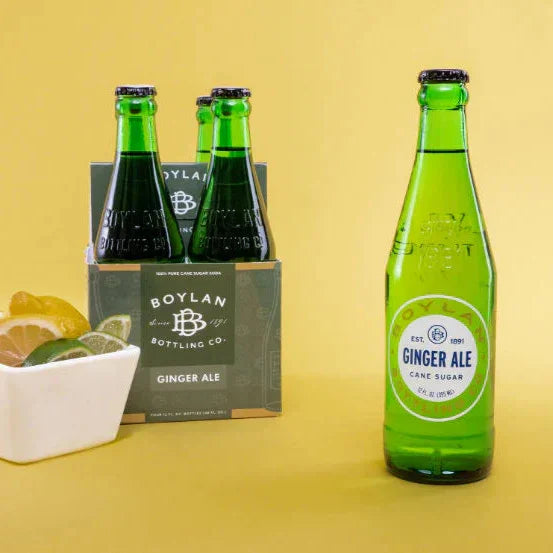 A 12oz bottle of Boylan Ginger Ale with a four-pack box in the background, displayed on a yellow backdrop, alongside a small bowl with lemon slices.