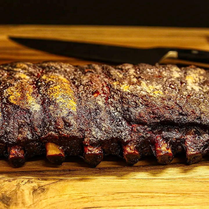 Two-thirds of a rack of Pen’s House ribs, glazed and sliced with bones disappearing out of frame and a large kitchen knife