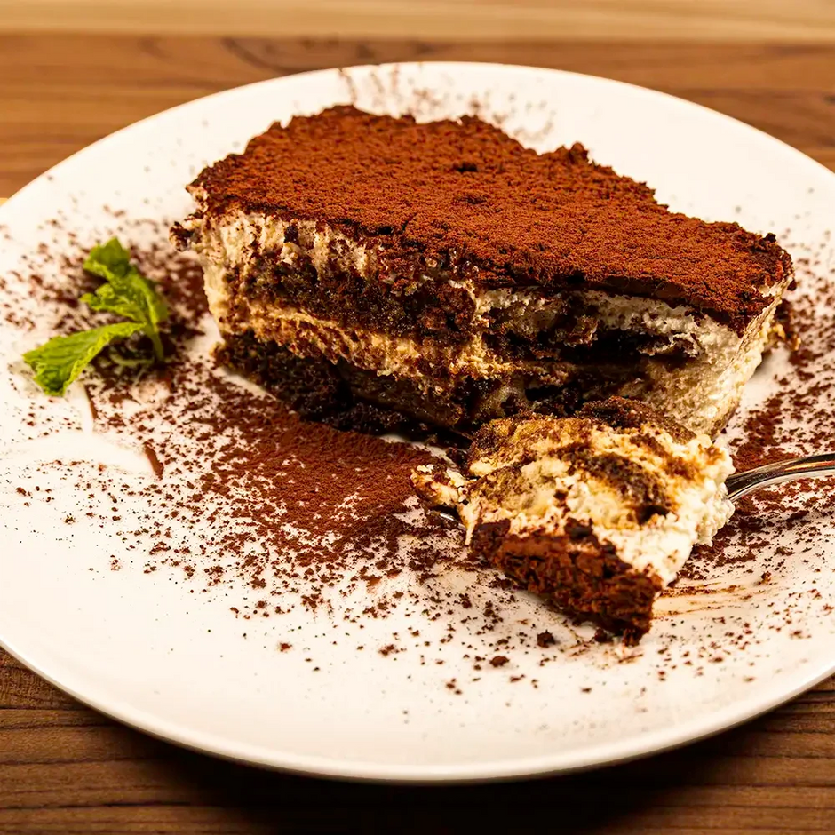 Slice of tiramisu on a white plate with cocoa powder and mint garnish.